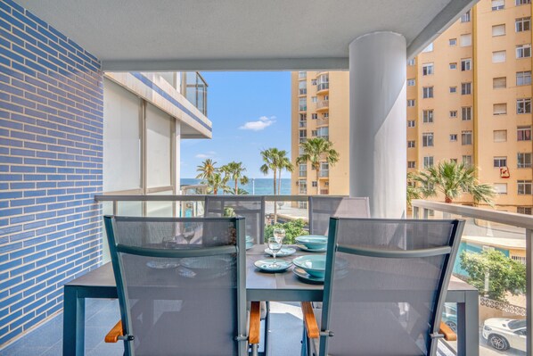A balcony with two chairs and a table, overlooking a cityscape with palm trees and a building with multiple windows.