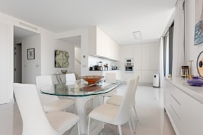 Een moderne keuken met witte kasten, een eettafel met glazen blad en witte stoelen.