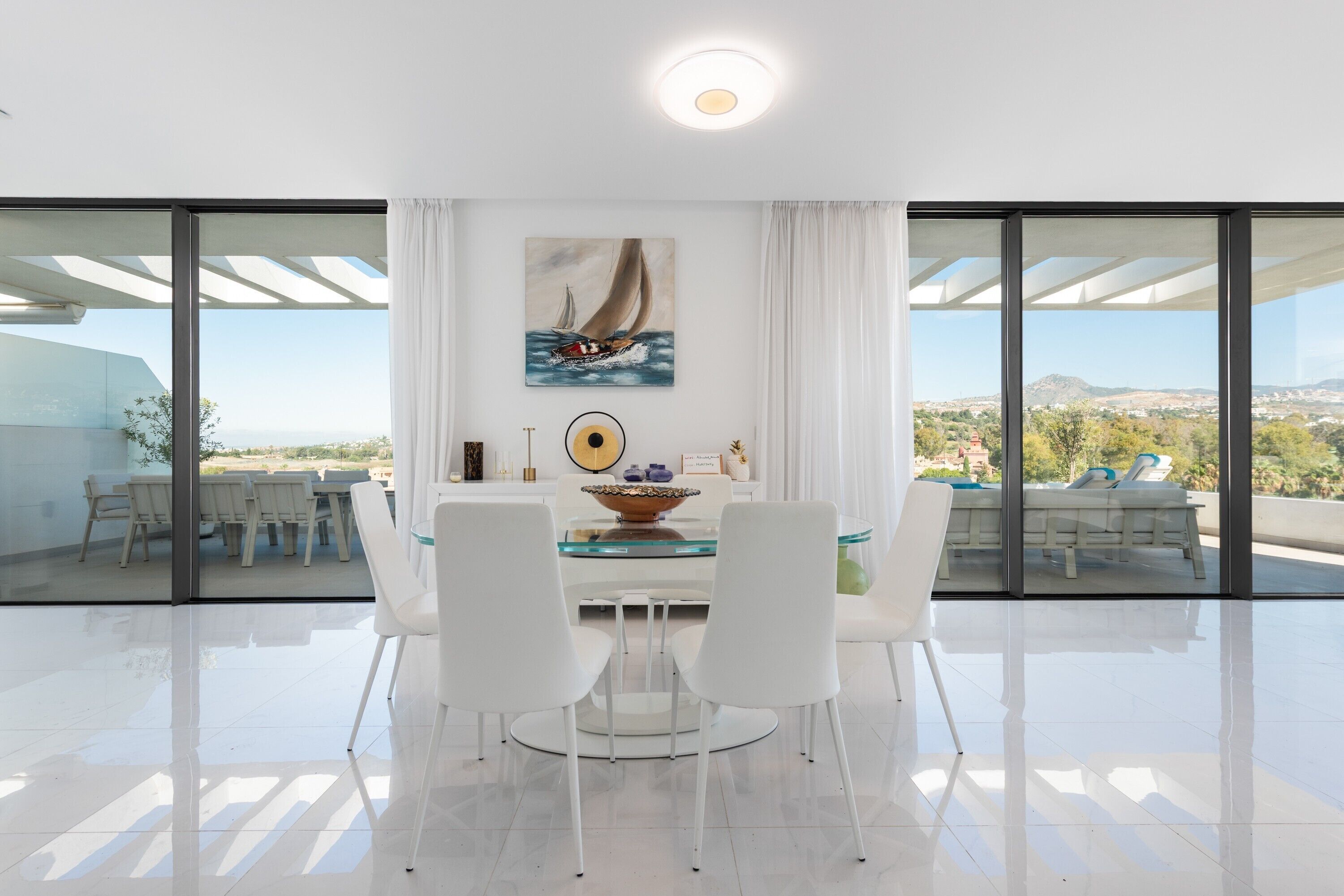 A modern dining area with a glass table, white chairs, a sailboat painting, and a view of the landscape through large windows.