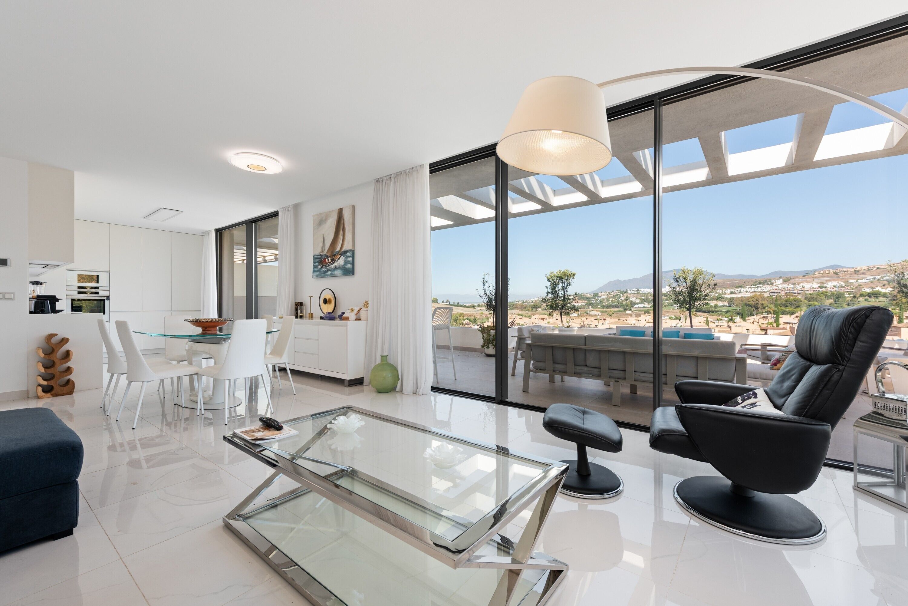 A modern living room with a glass coffee table, a black leather chair, and a dining area with white chairs.