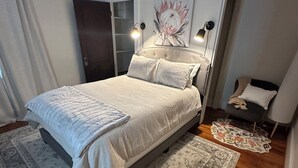 Ein Schlafzimmer mit einem Bett, einem Nachttisch, einem Stuhl, einem Gemälde und einem Fenster.