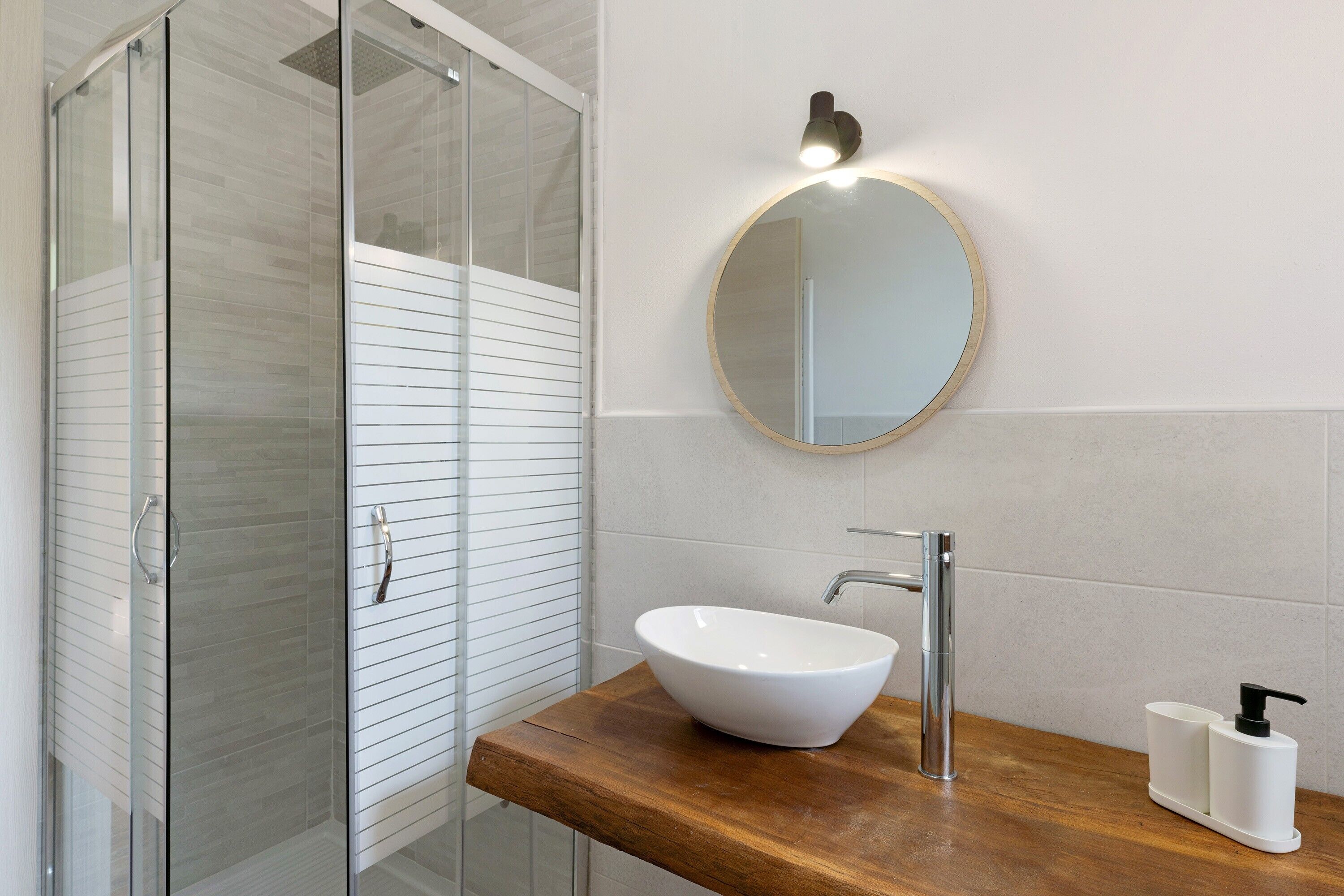 A bathroom with a glass shower enclosure, a white sink with a wooden countertop, a round mirror, and a wall-mounted light fixture.