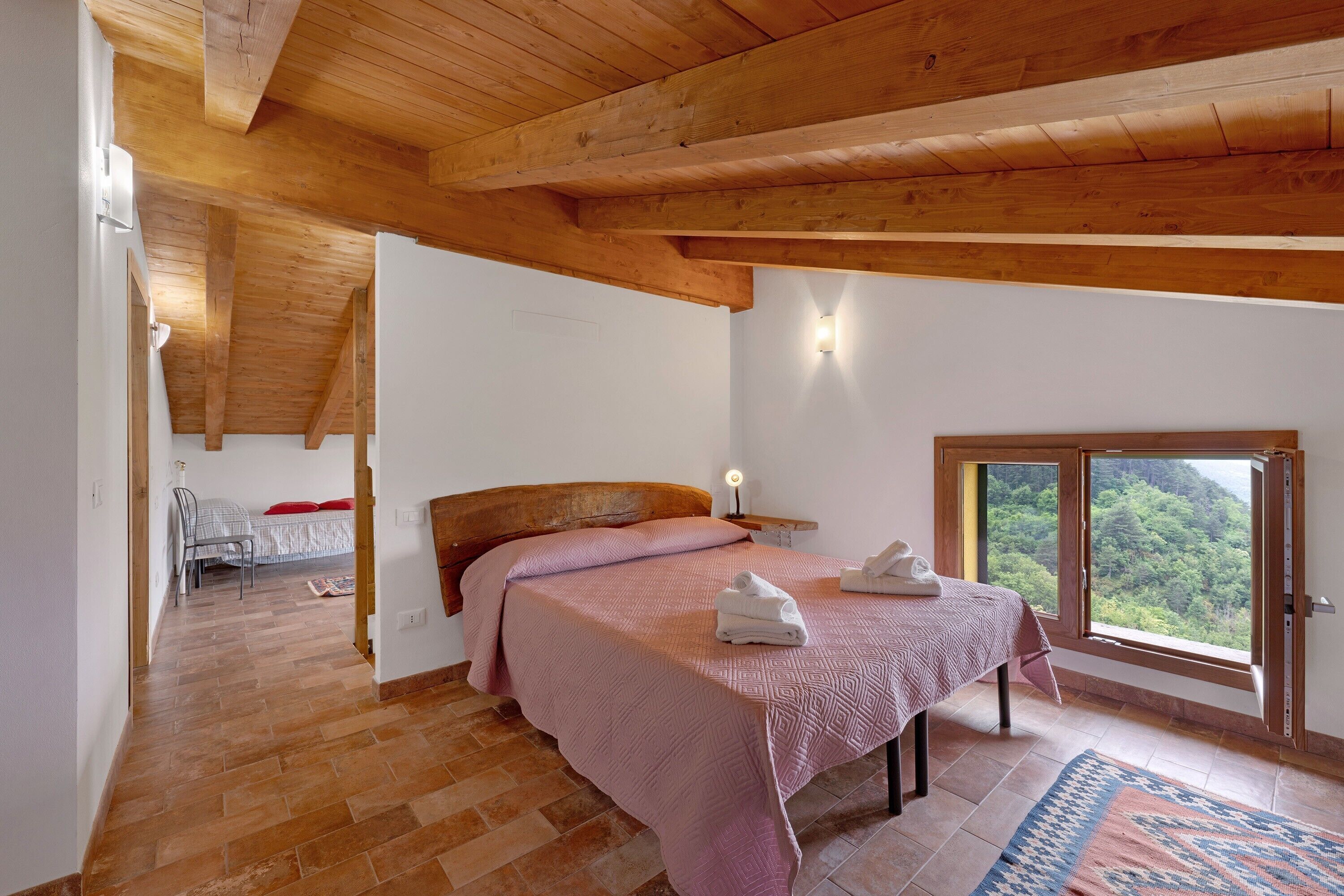 A bedroom with a wooden ceiling, a bed with a pink bedspread, a nightstand, a window with a view, and a wooden floor.