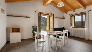 Habitación con techo de madera, paredes blancas, una mesa con sillas, un radiador y ventanas grandes con cortinas.