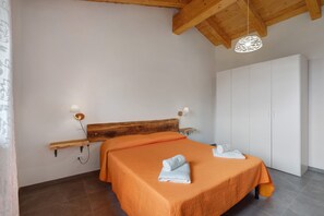 Un dormitorio con cabecera de madera, colcha naranja, dos toallas blancas, un armario blanco y una lámpara colgante.