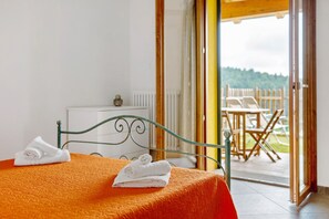 Ein Bett mit orangefarbener Bettdecke und zwei weißen Handtüchern, einem Metall-Kopfschild, einem weißen Heizkörper sowie Blick auf eine Au ßenpatio mit Stühlen und einem Holzzaun.