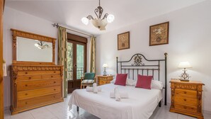A bedroom with a bed, wooden dresser, nightstands, a chandelier, and a door leading to a balcony.