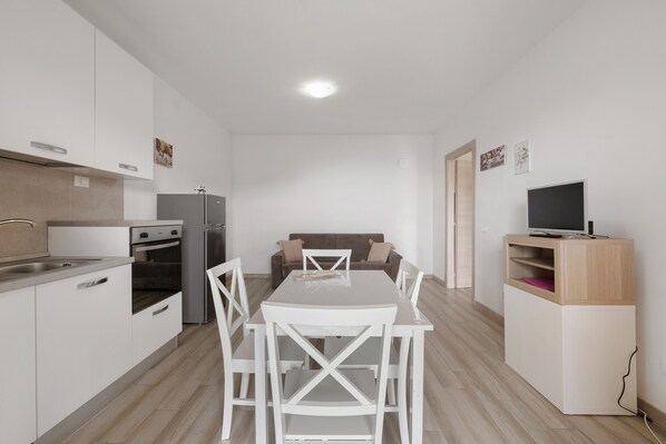 A modern kitchen with white cabinetry, a dining table with chairs, a sofa, and a TV on a stand.