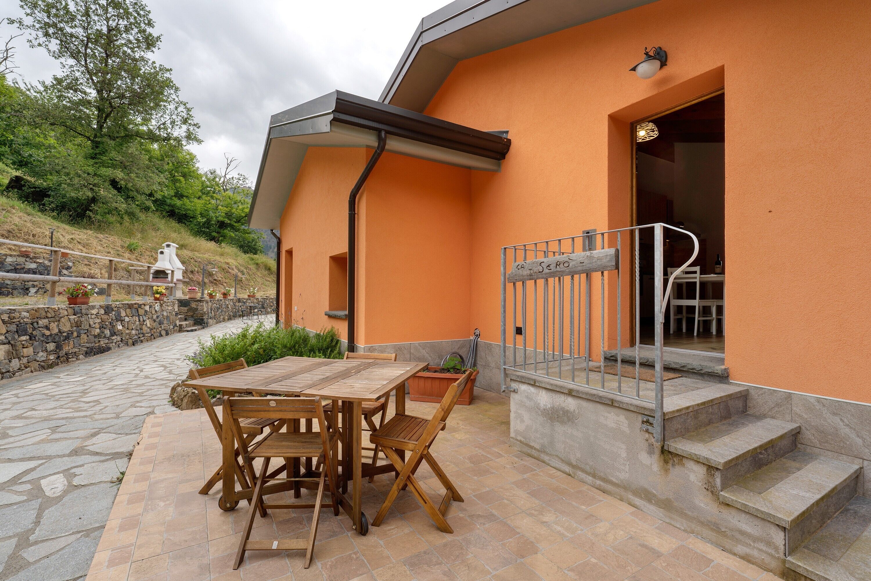 Een terras buiten met een houten tafel en stoelen, een stenen muur en een trap die naar een gebouw met een oranje gevel leidt.