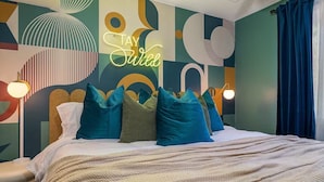 A modern bedroom with a geometric wall design, a neon sign that reads "Stay Wild", and a bed with multiple pillows. - Neon Dream Lodge! Pickleball, Hot Tub, Theater (McGaheysville)