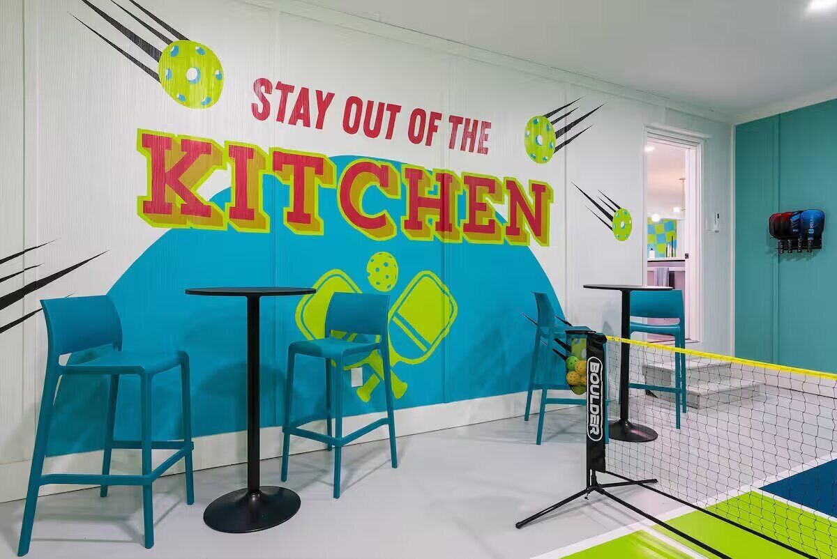 A brightly colored room with a wall mural saying "STAY OUT OF THE KITCHEN" and a netted area with chairs and tables.