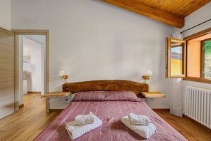 Quarto com cabeceira de madeira, cama com roupa de cama rosa, duas luminárias de mesa e janela com vista para o verde.