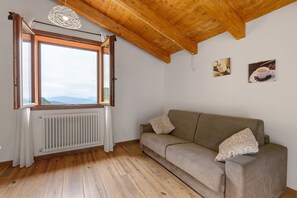 Um quarto com teto de madeira, janela com cortinas, radiador, sofá com almofadas estampadas e duas imagens penduradas na parede.