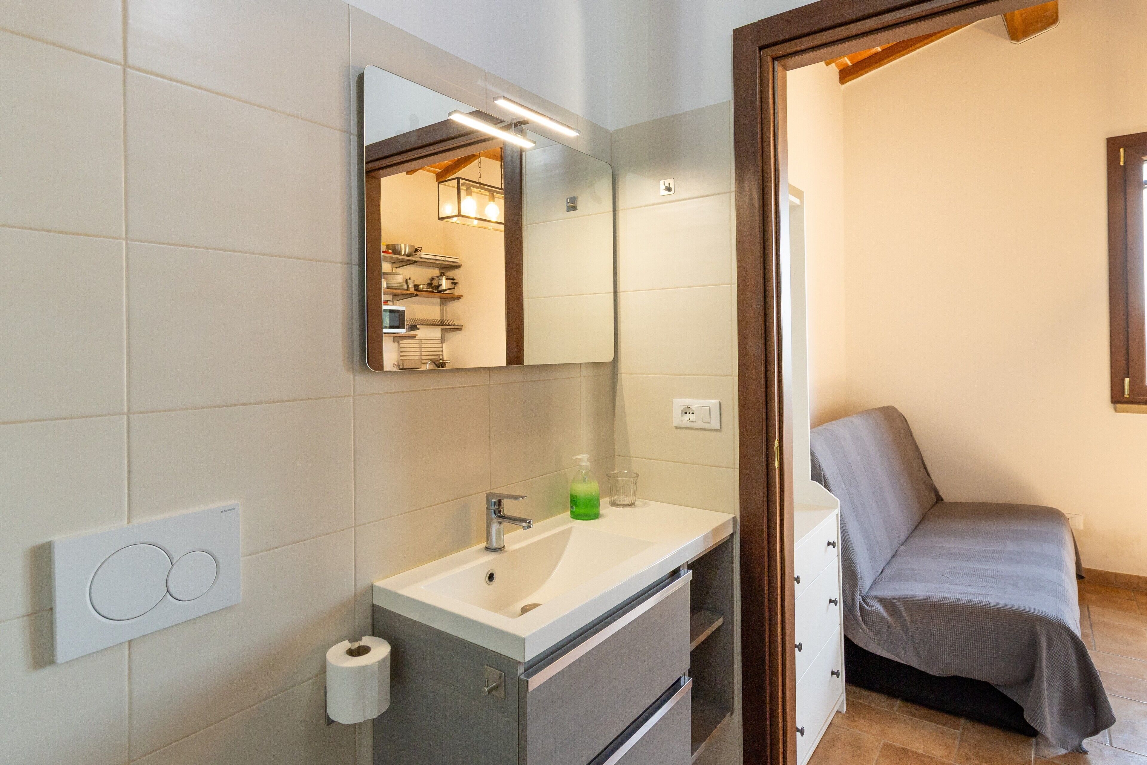 A bathroom with a sink, mirror, toilet paper holder, and a bed in the adjacent room.