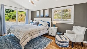 A bedroom with a bed, a nightstand, a chair, a window with a view of greenery, and a ceiling fan.