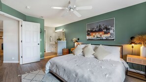 A bedroom with a bed, a ceiling fan, a nightstand, a lamp, and a cityscape painting on the wall.
