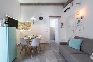A modern kitchen with a dining table, chairs, and a built-in fridge. There is a grey sofa with a teal pillow and a wall-mounted light fixture.