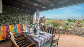 Un área de comedor al aire libre con una mesa preparada para cuatro personas, rodeada de sillas de madera, un muro de piedra y vistas al paisaje.
