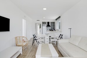 A modern, minimalist living room with a white sofa, a dining table with chairs, a TV mounted on the wall, and a kitchen area in the background.