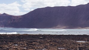 A rocky beach with waves, a person walking, and mountains in the background.