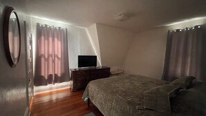 Une chambre à coucher avec un lit, une commode, un téléviseur posé sur la commode et une fenêtre avec des rideaux.