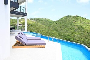 A rooftop infinity pool with lounge chairs, a dining area, and a view of a green hillside. - Villa Jasiri (Gros Islet)