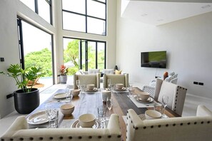 A dining area with a large table set for six, surrounded by chairs, with a TV mounted on the wall and a view of greenery outside. - Villa Jasiri (Gros Islet)