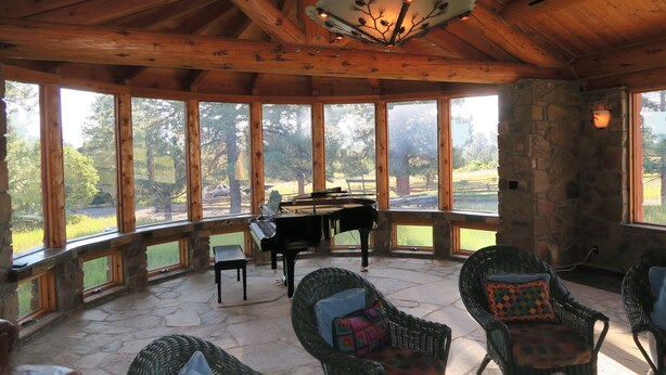 A covered outdoor area with stone walls, large windows, wicker furniture, and a grand piano. - Pine Grove Estate: Beautiful Log home on 40 acres with fantastic Mountain Views (Ridgway)