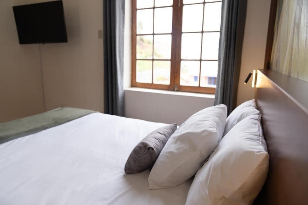 A neatly made bed with white and gray pillows, a flat-screen TV mounted on the wall, and a window with curtains.
