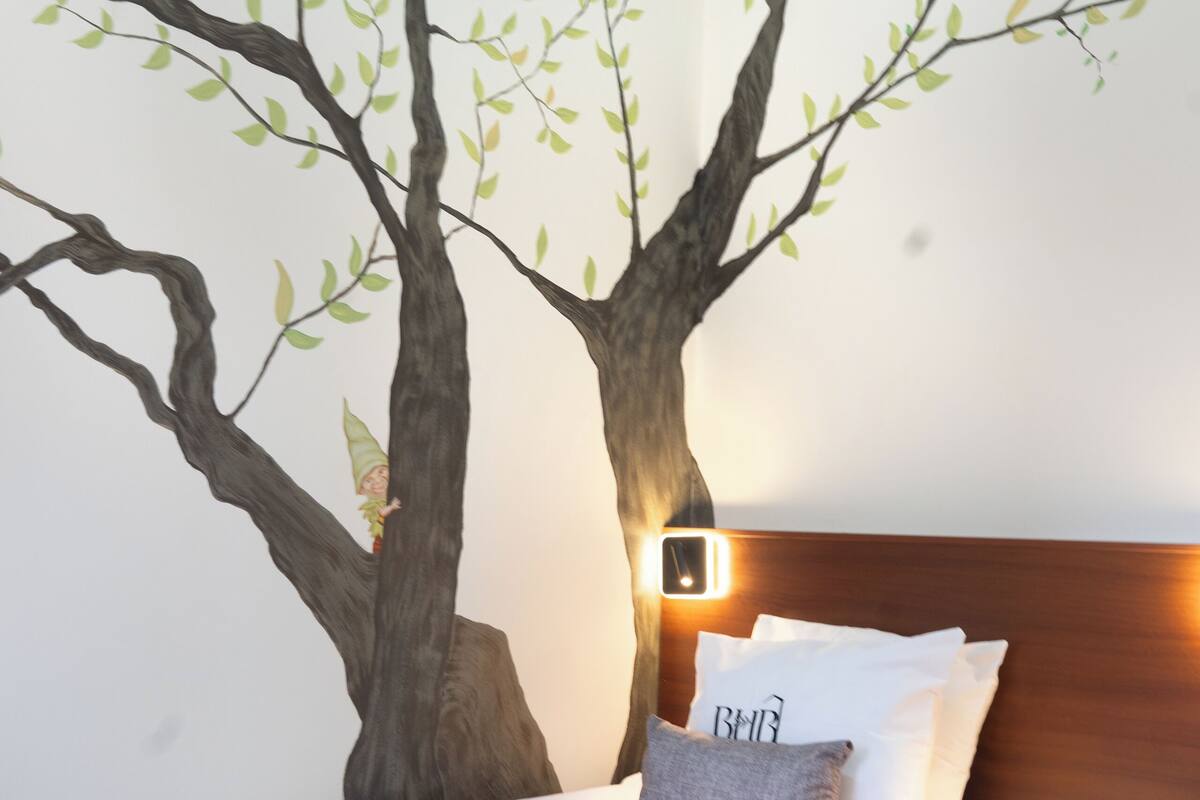 A bedroom with a bed, a nightstand, a wall-mounted lamp, and a tree mural.