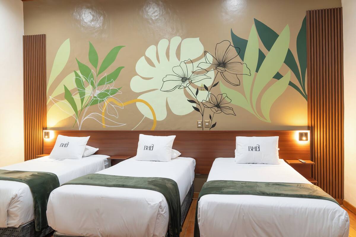 A hotel room with three single beds, each with a white bedspread and a pillow with the logo "BMB". The room features a large wall mural with floral designs and two wall-mounted reading lamps.