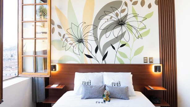A hotel room with a large bed, two pillows with "BMB" on them, a nightstand with a remote control, and a wall mural of flowers. - Bosco Hotel Boutique (Cusco)