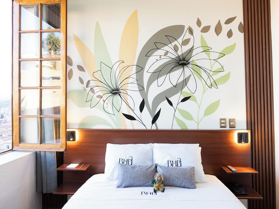 A hotel room with a large bed, two pillows with "BMB" on them, a nightstand with a remote control, and a wall mural of flowers.