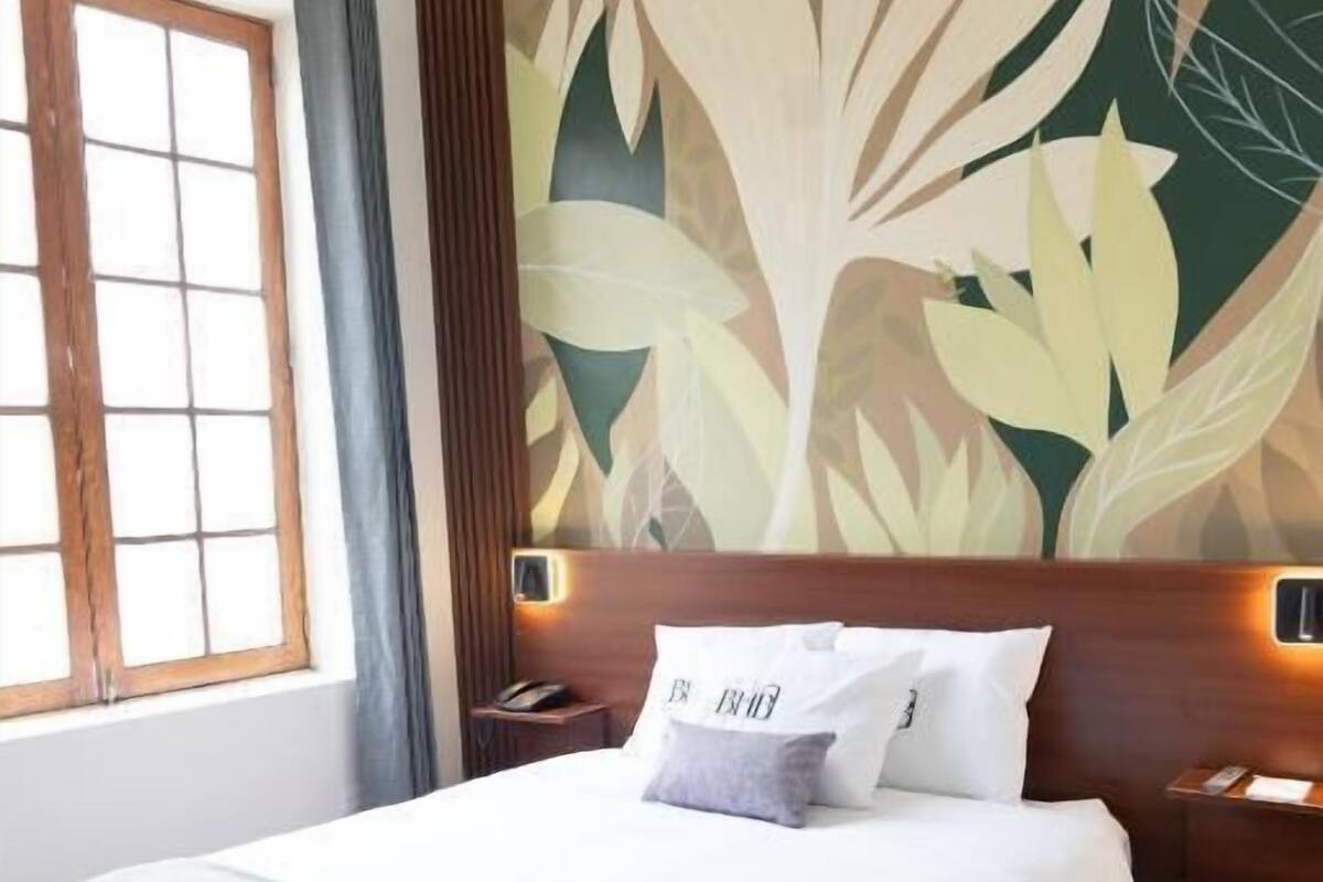 A neatly made bed with white linens and a green throw, a wooden headboard, and a wall with a large floral mural.