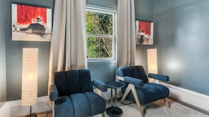 Un salon moderne avec deux fauteuils bleus, une petite table ronde et deux lampadaires. Il y a une fenêtre avec des rideaux et deux tableaux accrochés aux murs.