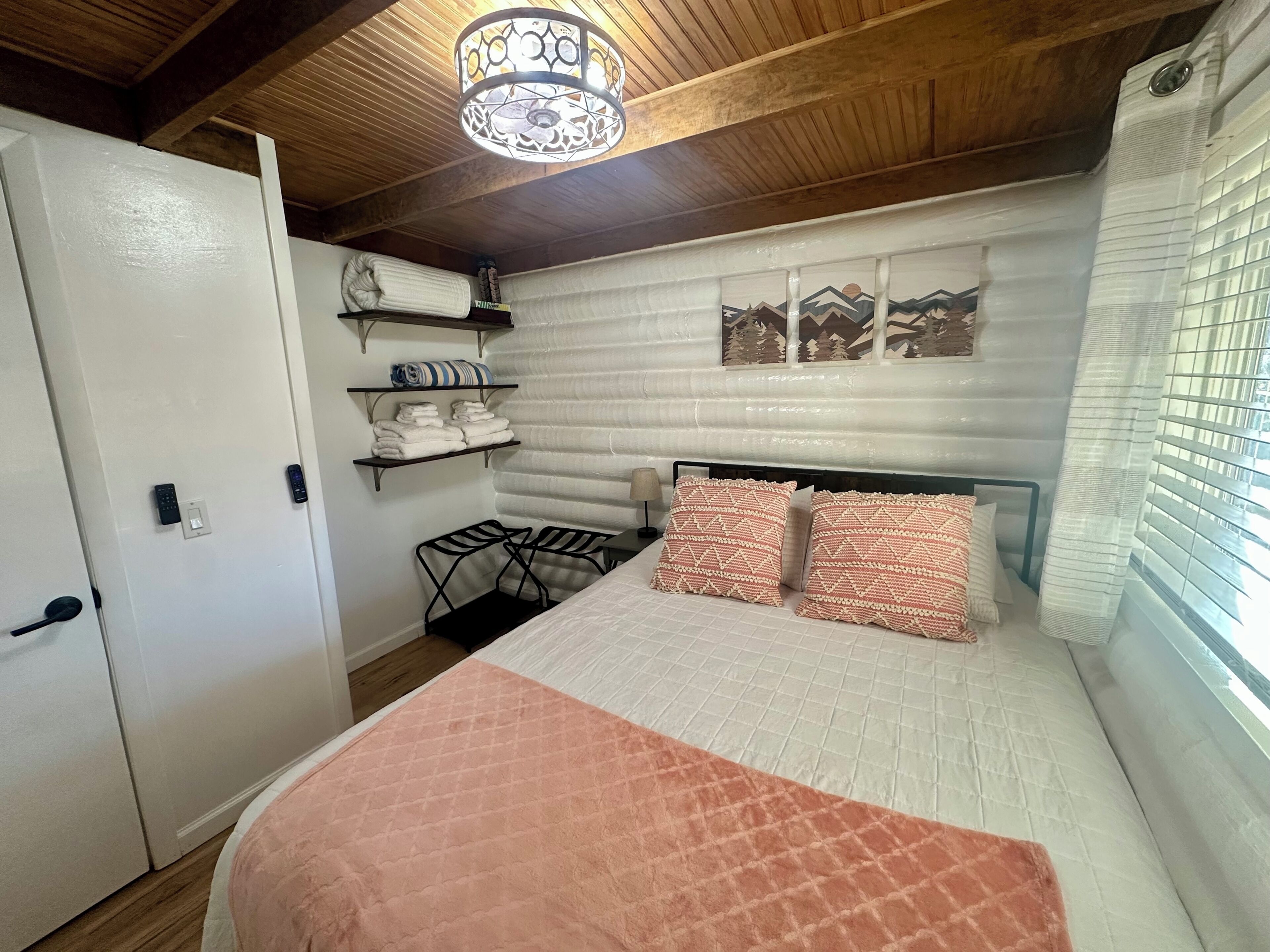 Ein Schlafzimmer mit Bett, einer Kommode, einem Regal mit Handtüchern, einem Sessel, einer Wandablage, einem Wandbild und einem Fenster mit Jalousien.