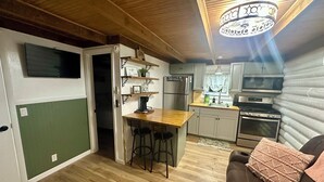 A compact kitchen with a wooden countertop, a refrigerator, a microwave, and a stove. There is a small dining area with stools and a coffee machine. The room has a green accent wall and a ceiling with a decorative light fixture.