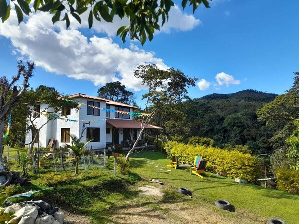 A white house with a red roof, surrounded by a lush green garden and a hilly landscape.