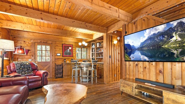 A cozy log cabin interior with a wooden dining area, leather seating, and a large flat-screen TV displaying a mountain lake.