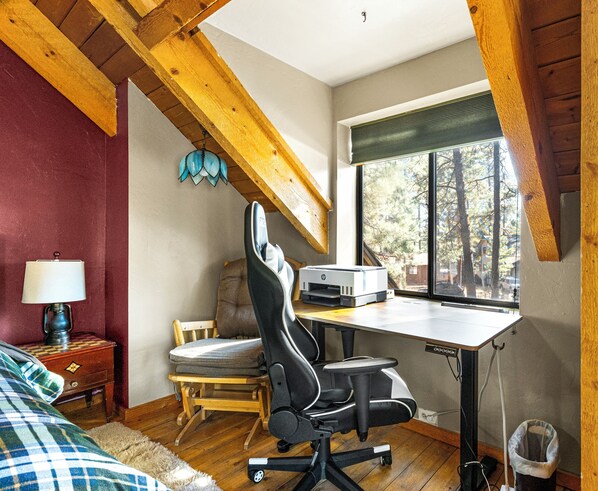A bedroom with a wooden ceiling, a desk with a printer, a chair, a lamp, and a window with a view of trees.