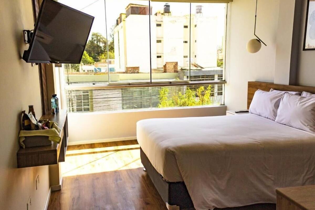 Un dormitorio con cama, televisor montado en la pared, ventana con vista a edificios y piso de madera.