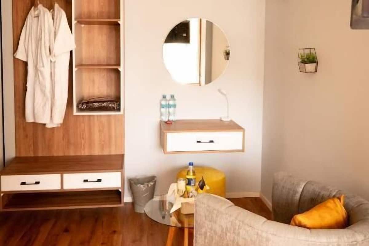 Una habitación de hotel moderna con un armario de madera, un espejo redondo, una pequeña mesa redonda con una silla amarilla y un sofá gris con un cojín amarillo.