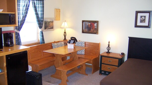 A compact room with a wooden bench, a small table, a bedside table with drawers, a lamp, a framed picture on the wall, and a bed. - Cozy Campground Cabin Cottage with WiFi and AC in charming Greybull (Greybull)