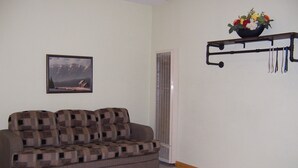 A living room with a sofa, a picture on the wall, a vent, and a shelf with a flower arrangement. - Cozy Campground Cabin Cottage with WiFi and AC in charming Greybull (Greybull)