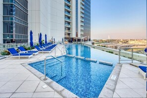 A rooftop pool with blue tiles, surrounded by lounge chairs and umbrellas, overlooking a cityscape. - Lux 2BR : Breathtaking Sea Views & Modern Comfort (Media City)
