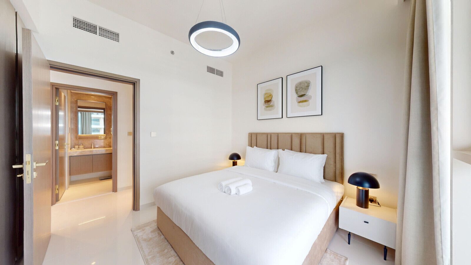 Ett hotellrum med en stor säng, sängbord, en modern lampa och ett badrum som syns genom en öppen dörr.
