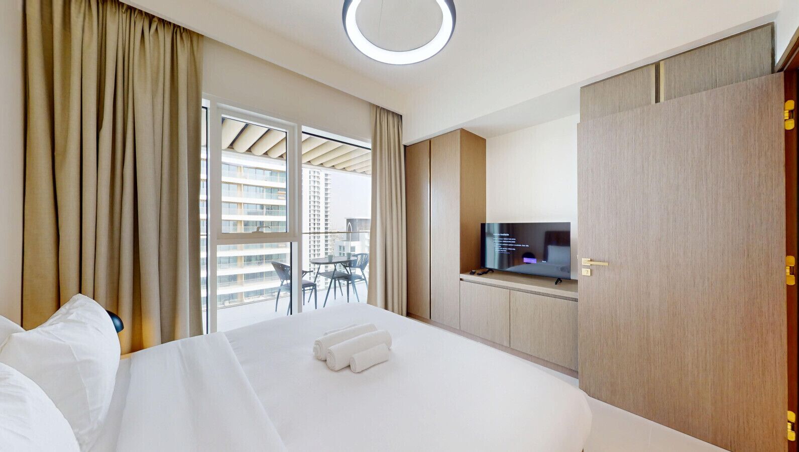 A hotel room with a large bed, a TV on a wooden cabinet, a balcony with a glass table and chairs, and a view of tall buildings.
