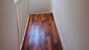 Una escalera estrecha con piso de madera y barandal blanco, que lleva a una habitación con un ventanal pequeño y una lámpara en el techo.