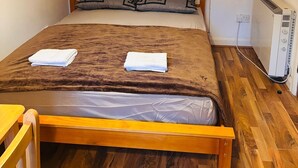 Una cama individual con cabecera de madera, dos almohadas, una manta marrón y una toalla blanca. Hay un calentador blanco en el suelo junto a la cama.