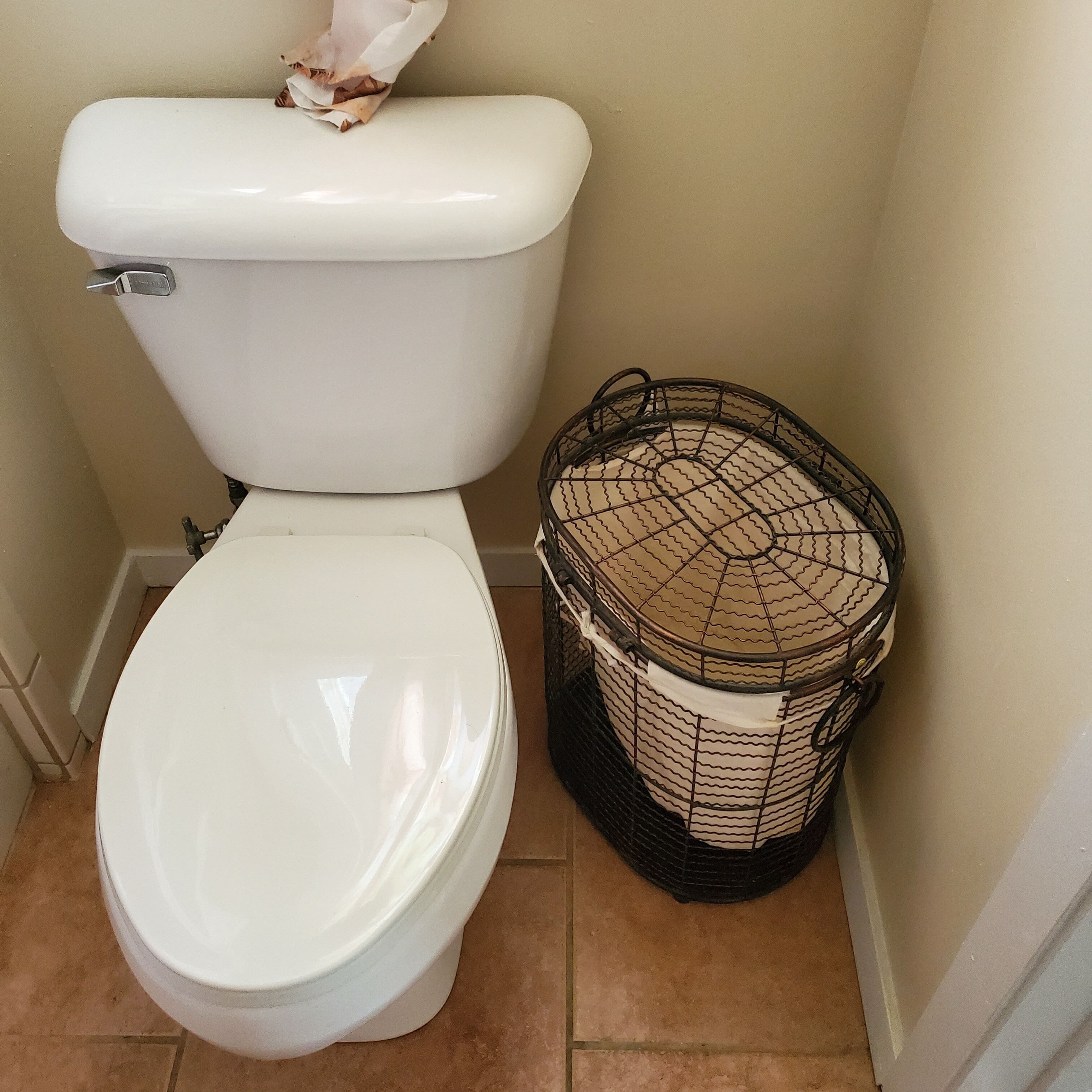 Des toilettes avec un abattant et un couvercle blancs, une cuvette en métal et un panier en fil de fer noir avec un couvercle blanc placé à côté.